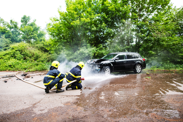 Two firefighters extinguishing a burning car after an accident on the road in the countryside.