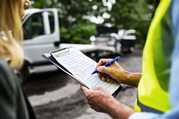 An unrecognizable man insurance agent talking to woman outside on the road after a car accident, making notes.