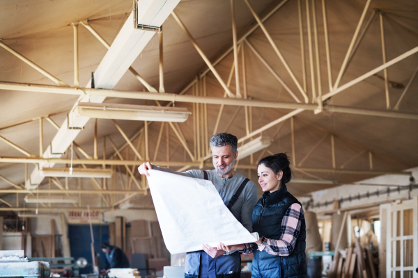 Portrait of a man and woman workers in the carpentry workshop, looking at paper plans.