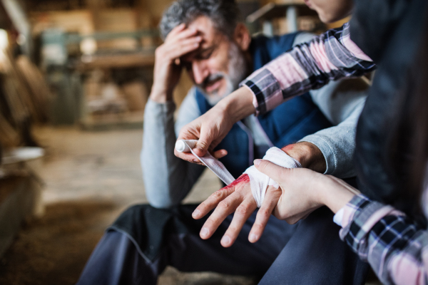 An unrecognizable woman bandaging a hand of a man worker after an accident in carpentry workshop. A woman helping her colleague at work.