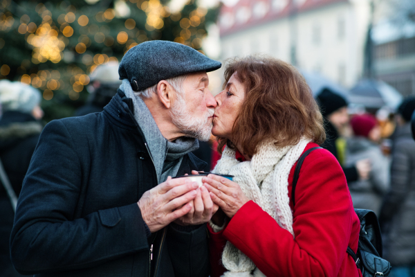 Happy senior couple on an outdoor Christmas market. Man and woman holding enameled cups, kissing. Winter time.