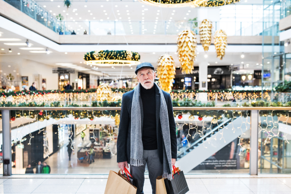 A portrait of bored and tired senior man with paper bags standing in shopping centre at Christmas time.