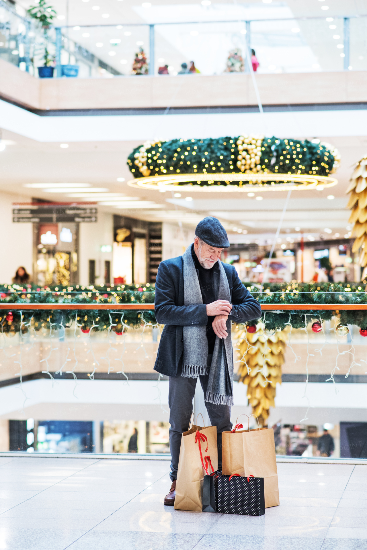 A senior man with bags standing in a shopping center at Christmas time, checking the time.