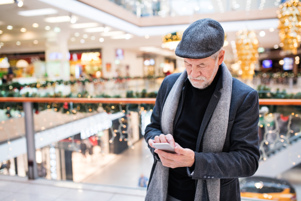 Senior man with smartphone doing Christmas shopping. Man text messaging. Shopping center at Christmas time.