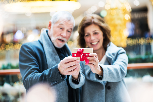 A portrait of senior couple holding a wrapped present in front of them in shopping center at Christmas time.