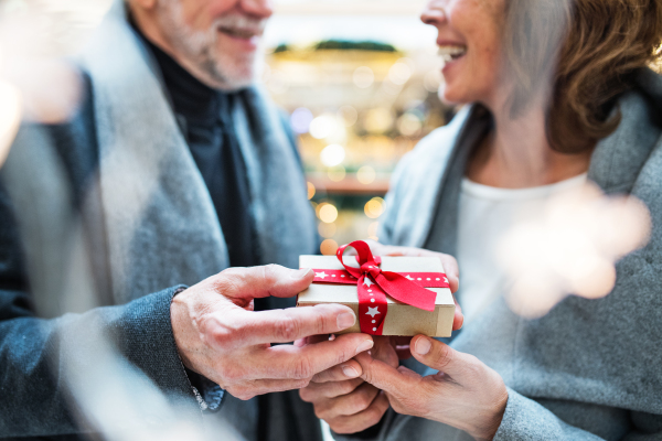 An unrecognizable senior couple holding a wrapped present in front of them in shopping center at Christmas time.