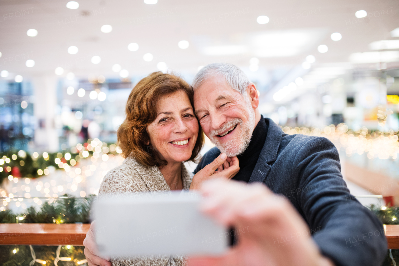 Senior couple doing Christmas shopping. Man and woman with smartphone taking a selfie. Shopping center at Christmas time.