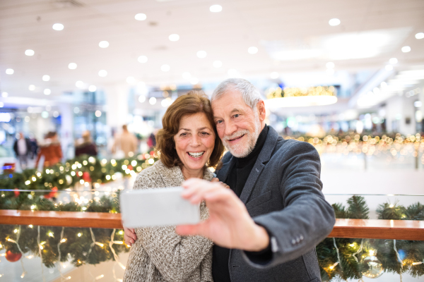 Happy senior couple with smartphone taking selfie in shopping center at Christmas time.