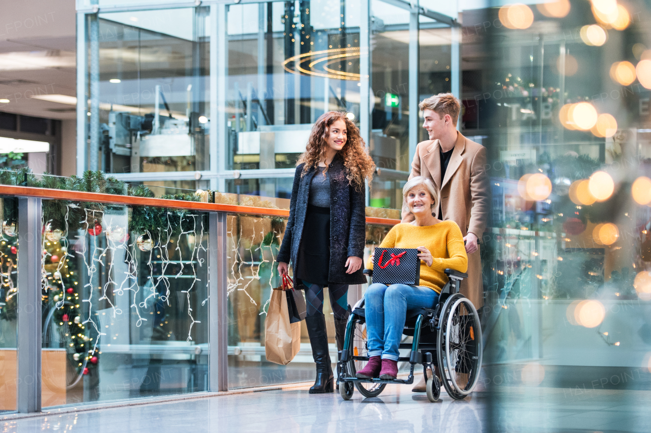 A senior grandmother in wheelchair and teenage grandchildren with paper bags walking in shopping center at Christmas time.