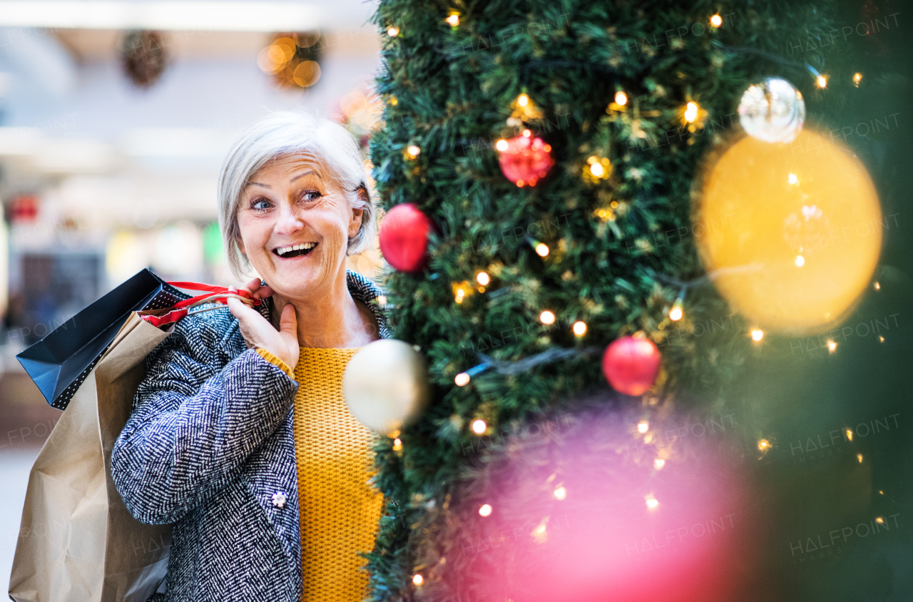 A portrait of senior woman with paper bags in shopping center at Christmas, standing by a Christmas tree.