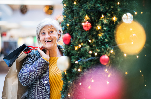 A portrait of senior woman with paper bags in shopping center at Christmas, standing by a Christmas tree.