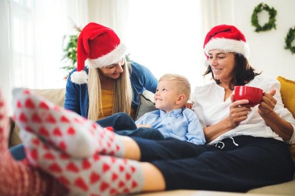 A small boy with mother and grandmother with Santa hat sitting on a sofa at home at Christmas time, talking.