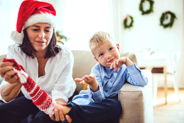 A small boy and grandmother with a Santa hat sitting on a sofa at home at Christmas time, putting on socks.