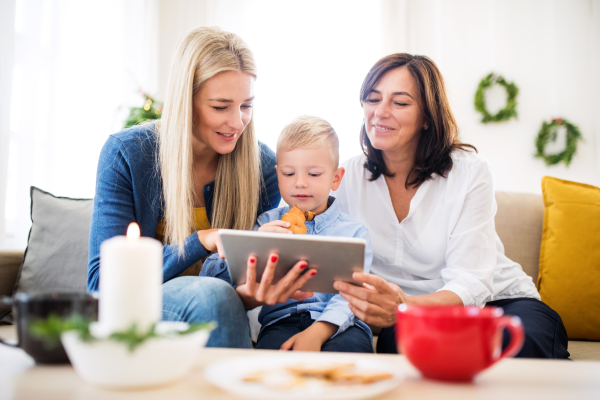 A small boy with mother and grandmother sitting on a sofa at home at Christmas time, using tablet.