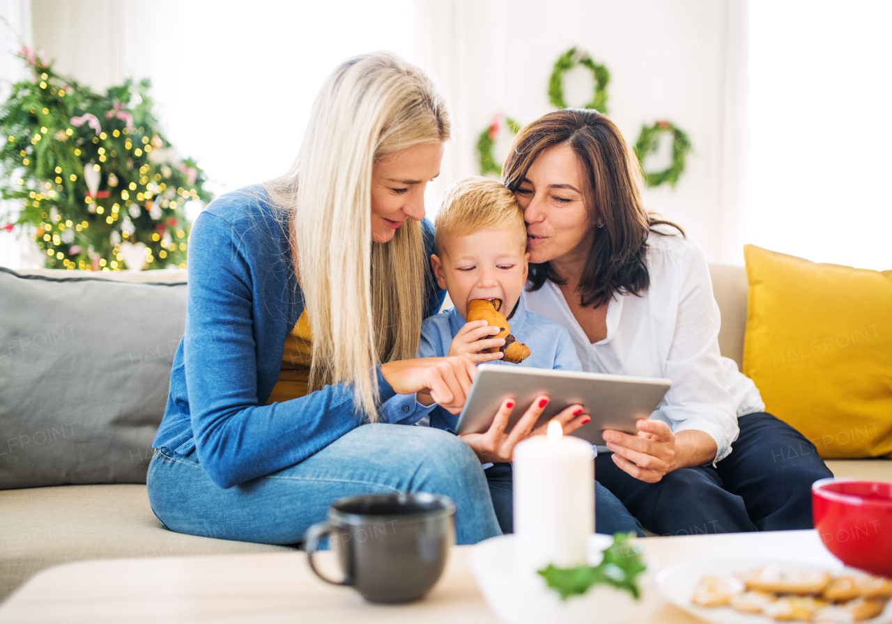 A small boy with mother and grandmother sitting on a sofa at home at Christmas time, taking selfie with tablet.