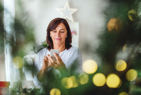 A senior woman with smartphone standing by a Christmas tree at Christmas time at home.