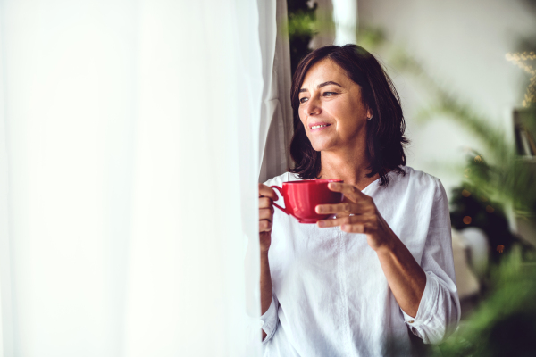 A senior woman with a cup standing by the window at home at Christmas time, looking out. Copy space.