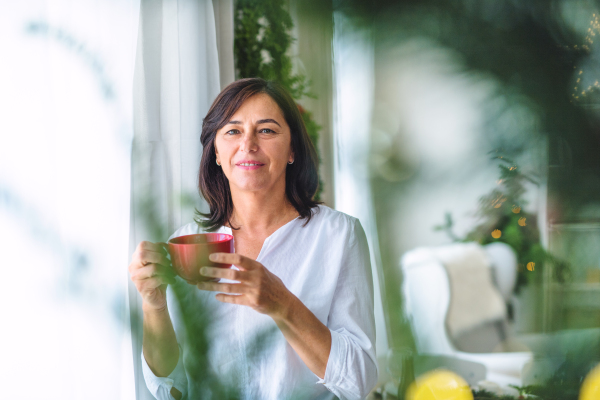 A senior woman with a cup standing by the window at home at Christmas time. Copy space.