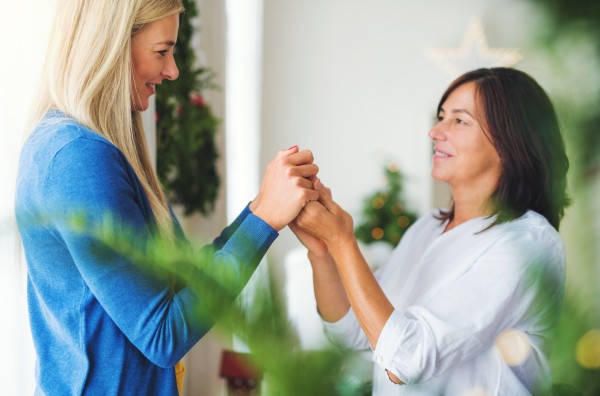 A happy senior woman with adult daughter standing by Christmas tree at home, holding hands.