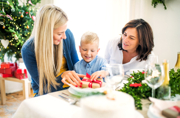 A small boy with present and mother and grandmother sitting at a table at home at Christmas time.