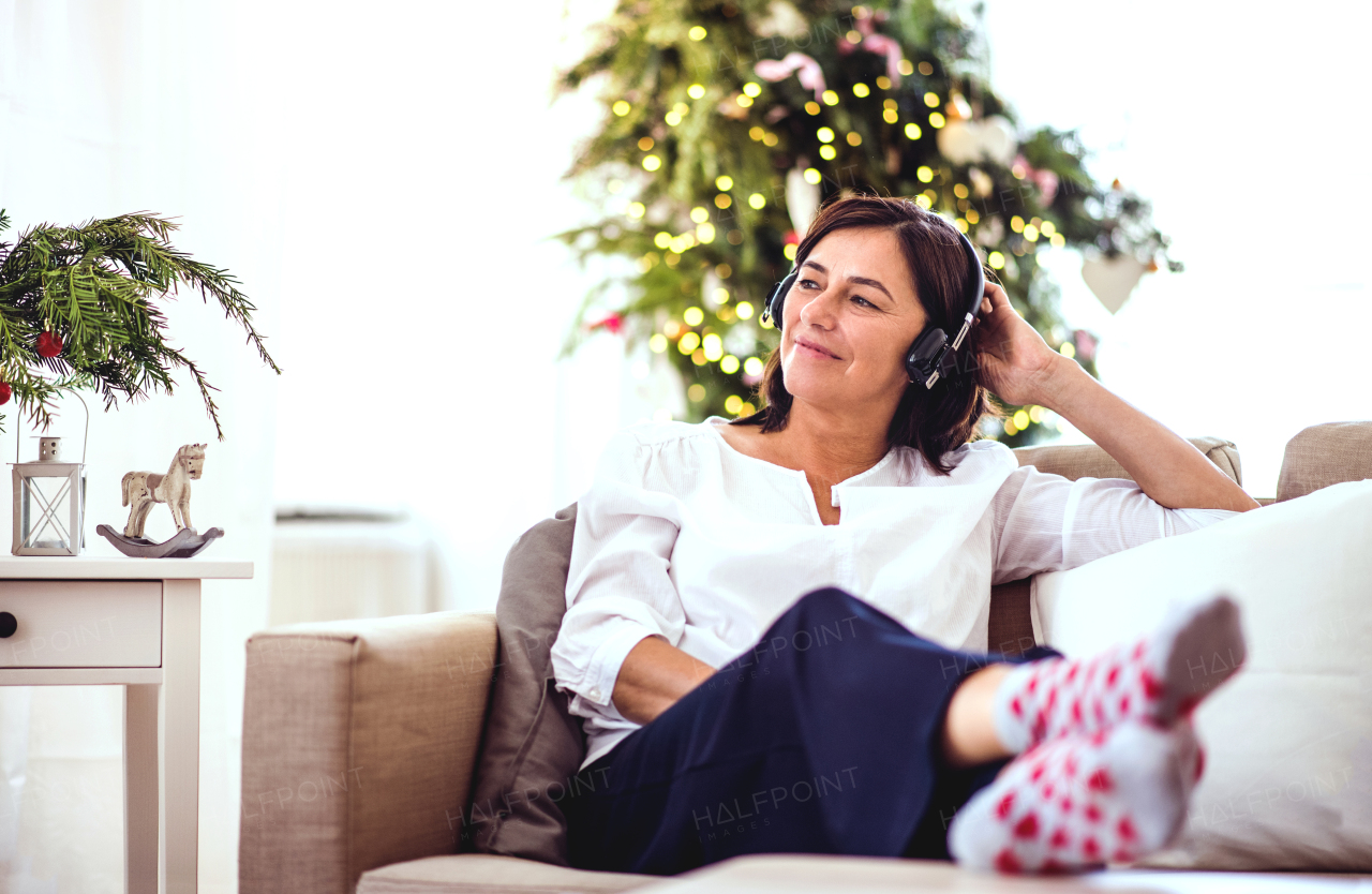 A senior woman with headphones and feet on a coffee table sitting on a sofa at home, listening to music at Christmas time.