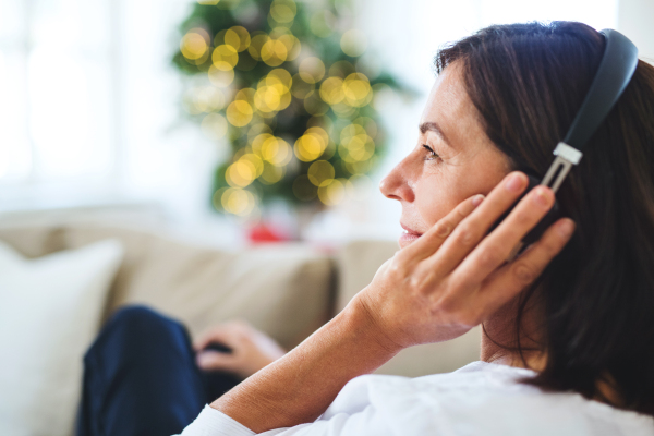 A senior woman with headphones sitting on a sofa at home, listening to music at Christmas time.