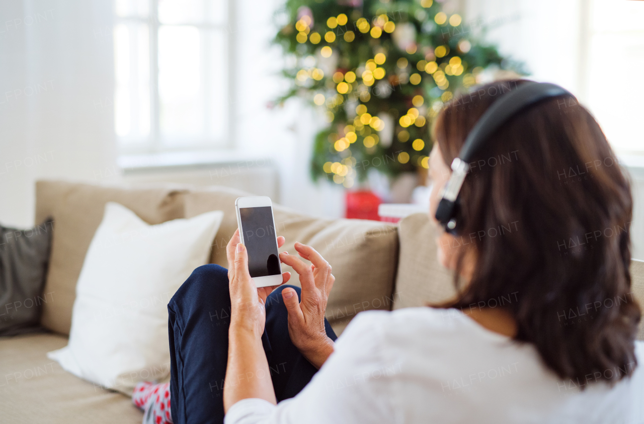 A senior woman with headphones and smartphone sitting on a sofa at home, listening to music at Christmas time. Rear view.