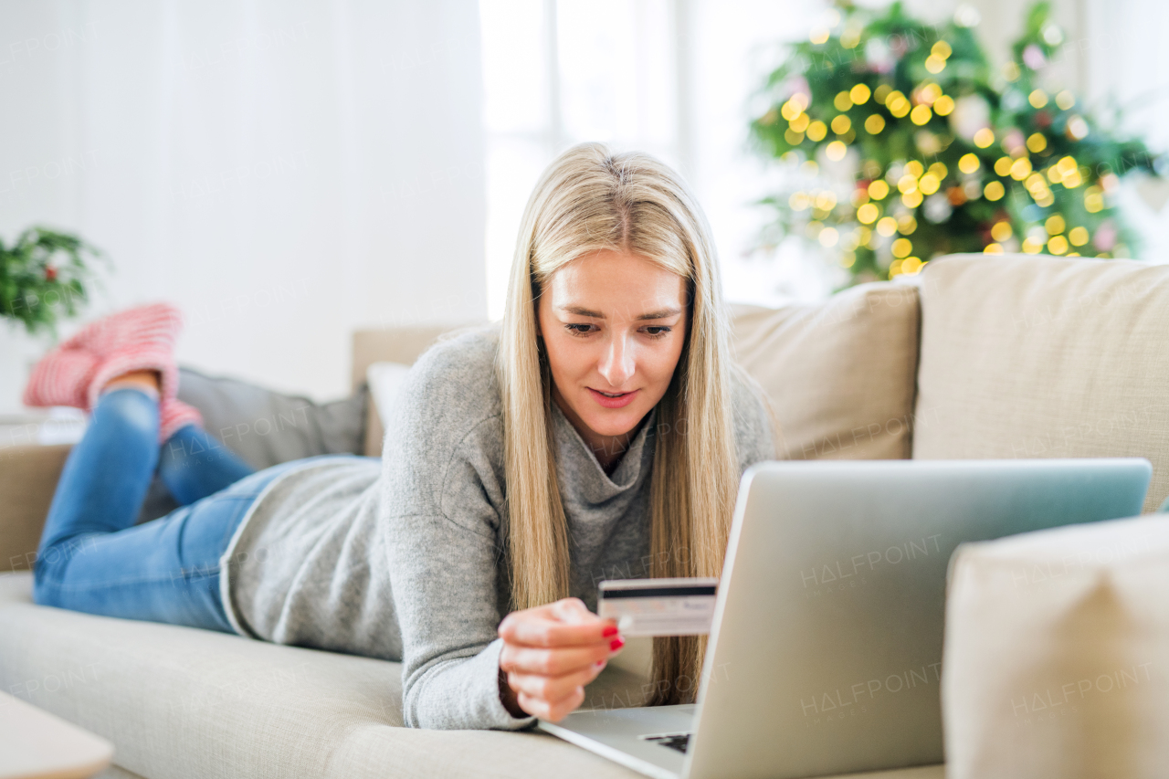 A young woman with credit card and laptop lying on a sofa at home at Christmas time, making online payment.