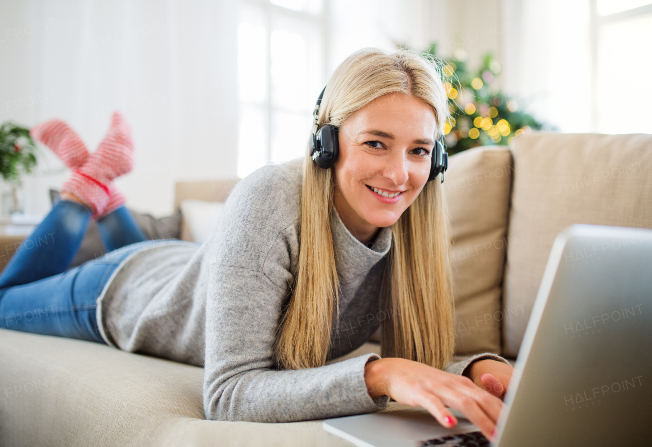 A young woman with headphones and laptop lying on a sofa at home at Christmas time.