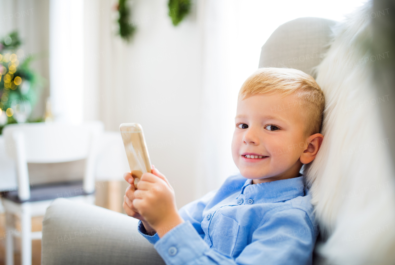 A small boy with smartphone sitting on an armchair at home at Christmas time, playing games.