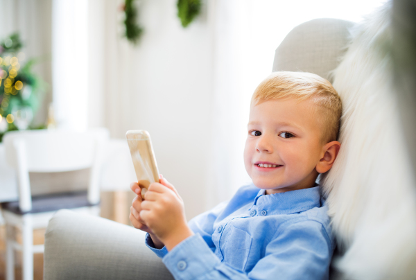 A small boy with smartphone sitting on an armchair at home at Christmas time, playing games.