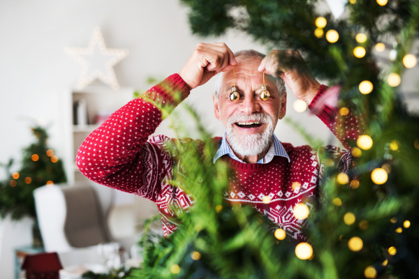 A cheerful senior man standing by Christmas tree, putting balls in front of his eyes.