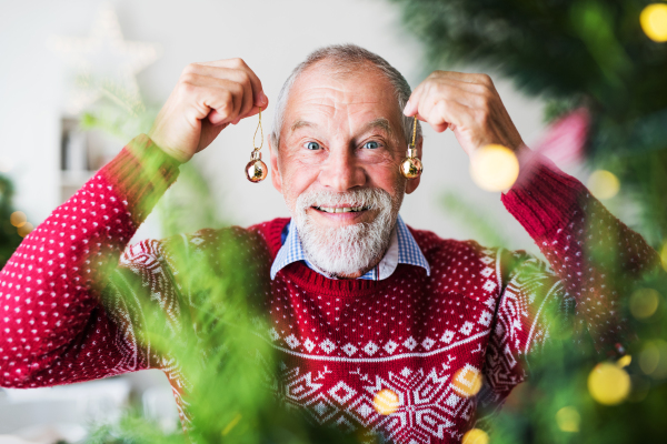 A cheerful senior man standing by Christmas tree, holding balls ornaments.