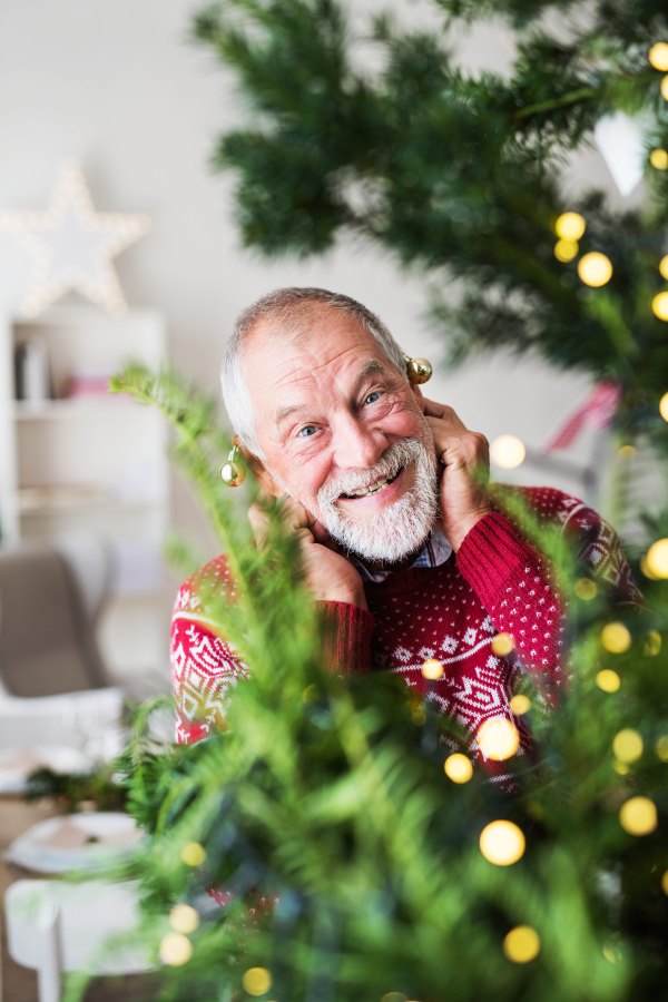 A cheerful senior man standing by Christmas tree, putting ball ornaments on his ears.