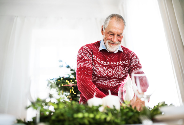 A happy senior man setting a table for a dinner at home at Christmas time.