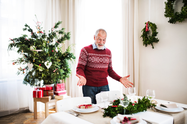 A surprised senior man standing by a table set for a dinner at home at Christmas time.