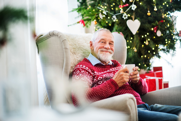 A portrait of senior man sitting on armchair at home at Christmas time, holding a white cup.