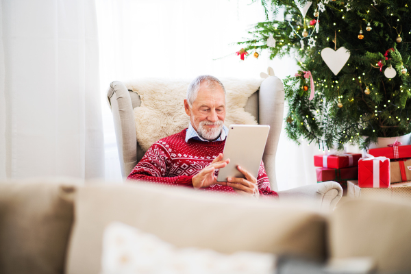 A portrait of senior man sitting on armchair at home at Christmas time, using tablet.