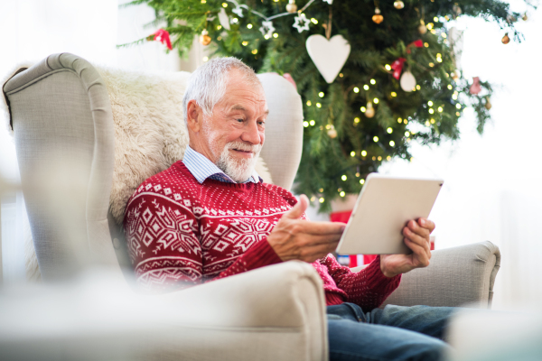 A portrait of senior man sitting on armchair at home at Christmas time, using tablet.