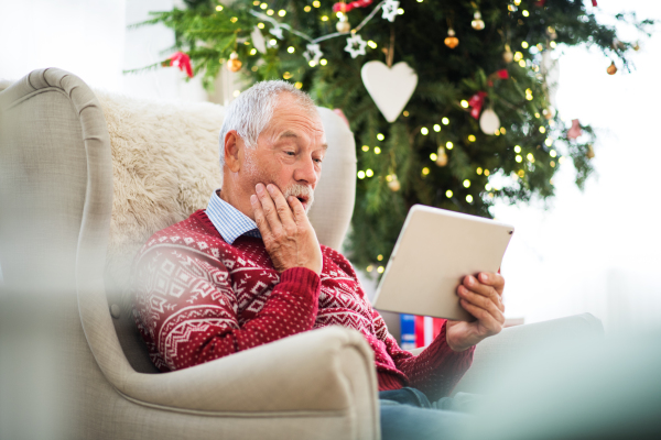 A surprised senior man sitting on an armchair by a Christmas tree, using tablet. Festive time.