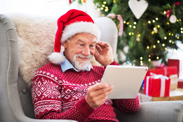 A portrait of senior man with Santa hat sitting on armchair at home at Christmas time, using tablet.