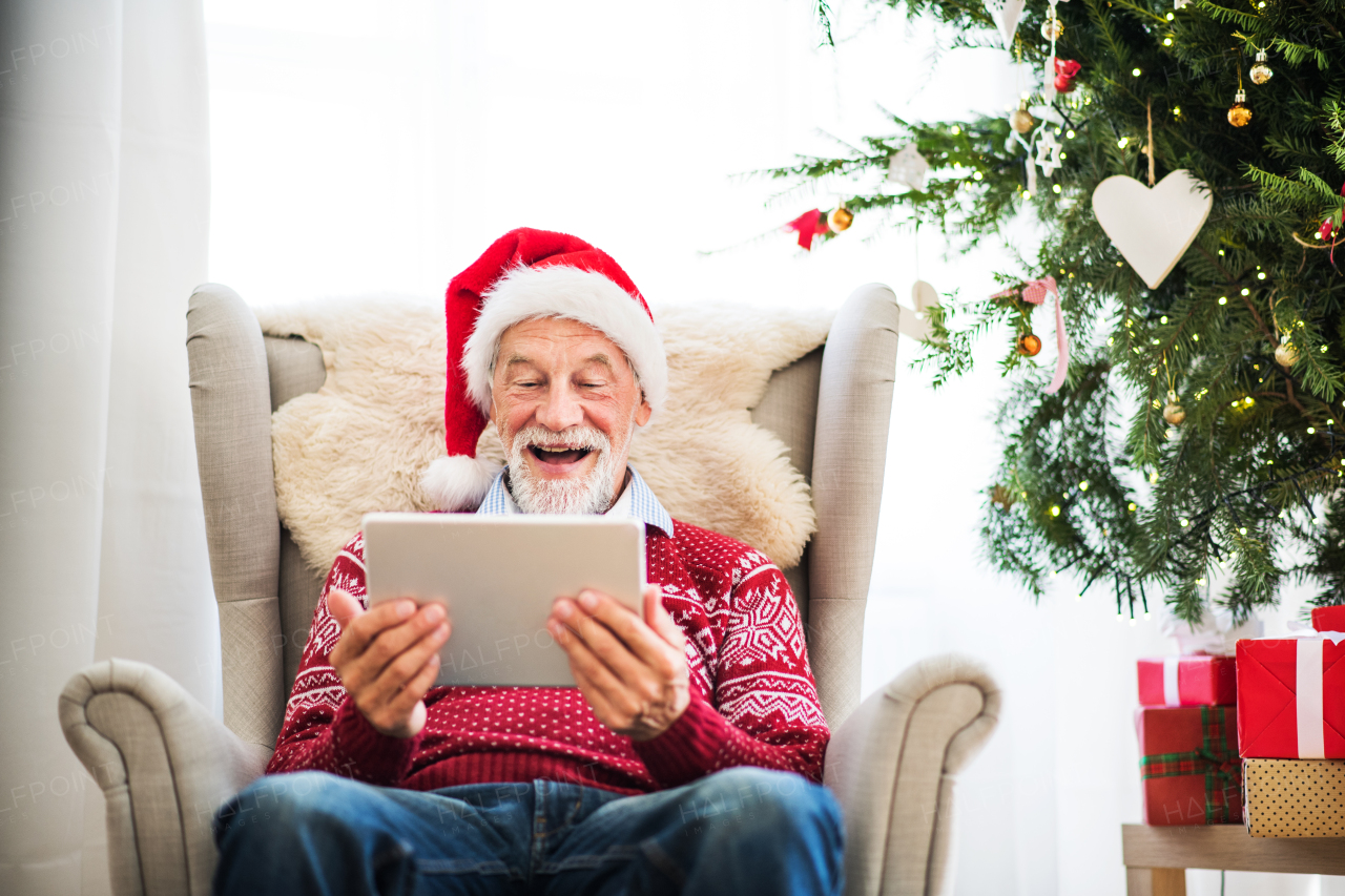 A portrait of senior man with Santa hat sitting on armchair at home at Christmas time, using tablet.