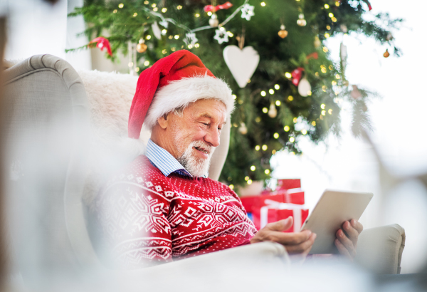 A portrait of senior man with Santa hat sitting on armchair at home at Christmas time, using tablet.