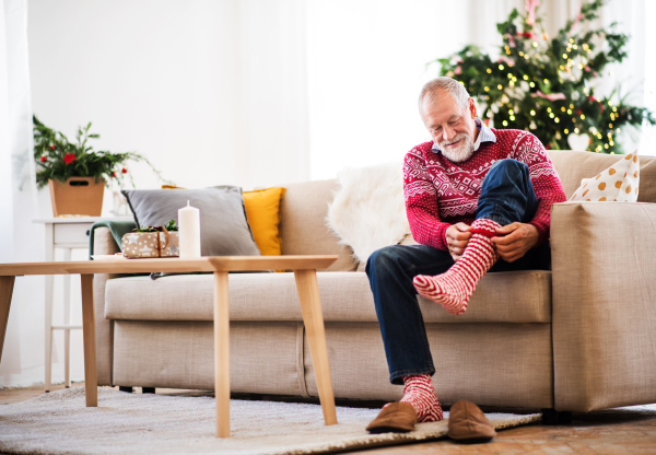 A senior man sitting on a sofa at home at Christmas time, putting socks on. Copy space.