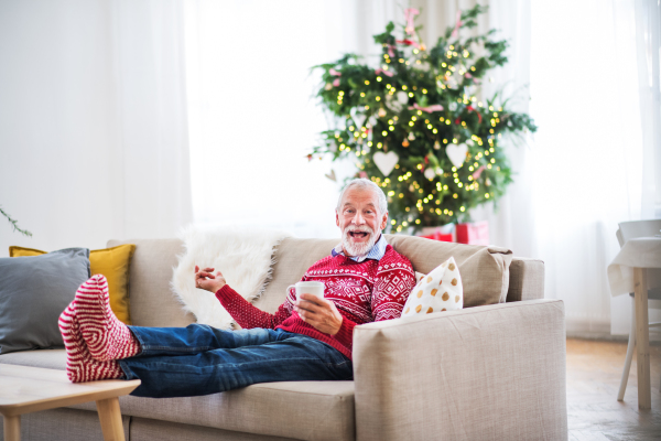 A senior man with feet on a coffee table sitting on a sofa at home, holding a cup of coffee at Christmas time.