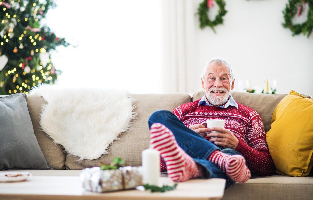 A senior man with feet on a coffee table sitting on a sofa at home, holding a cup of coffee at Christmas time.