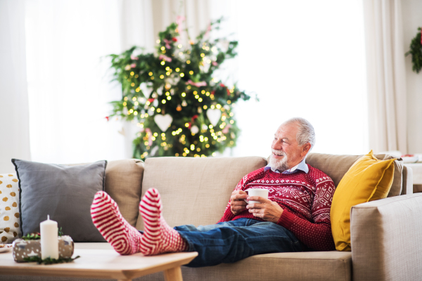 A senior man with feet on a coffee table sitting on a sofa at home, holding a cup of coffee at Christmas time.