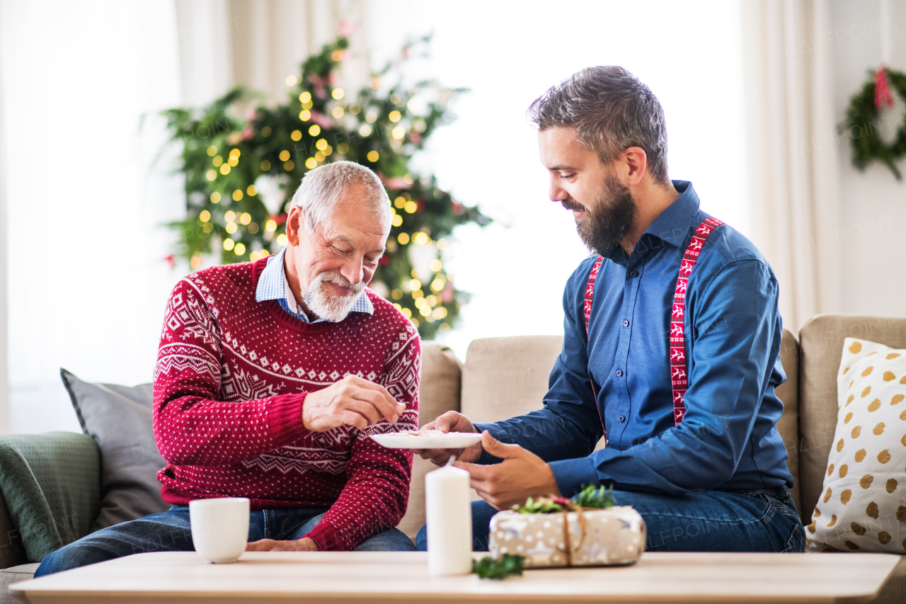 A senior father and adult son sitting on a sofa at home at Christmas time, eating biscuits and talking.