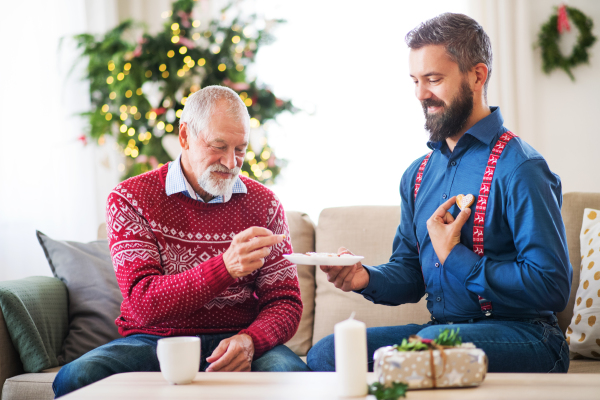 A senior father and adult son sitting on a sofa at home at Christmas time, eating biscuits and talking.