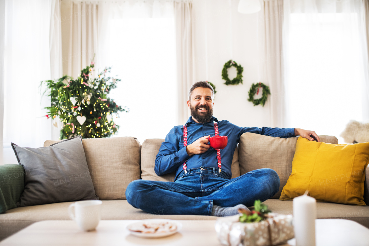 A ceherful hipster man with a red cup sitting on a sofa at home at Christmas time.